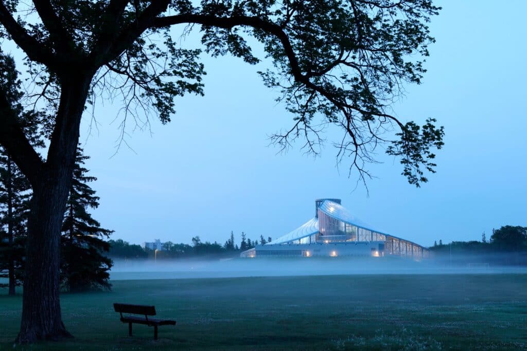 Long shot of The Leaf at Assiniboine Park at dusk, with the glowing building reflected in the mist across a vast green field.