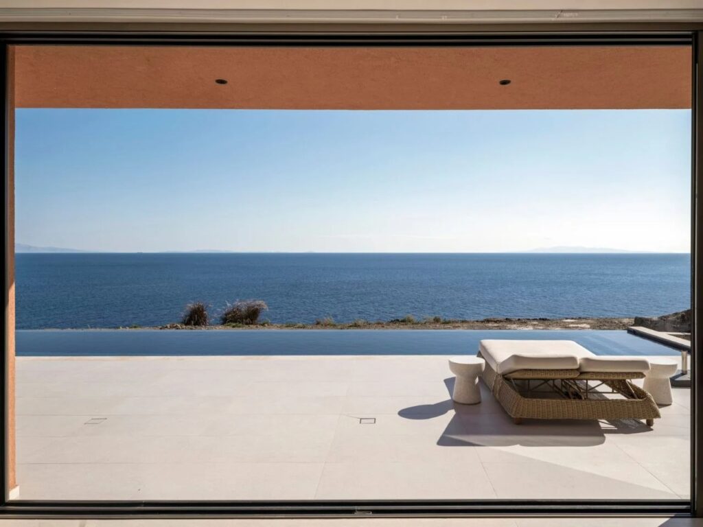 Minimalist sun loungers on a white stone terrace overlooking an infinity pool and the deep blue sea.