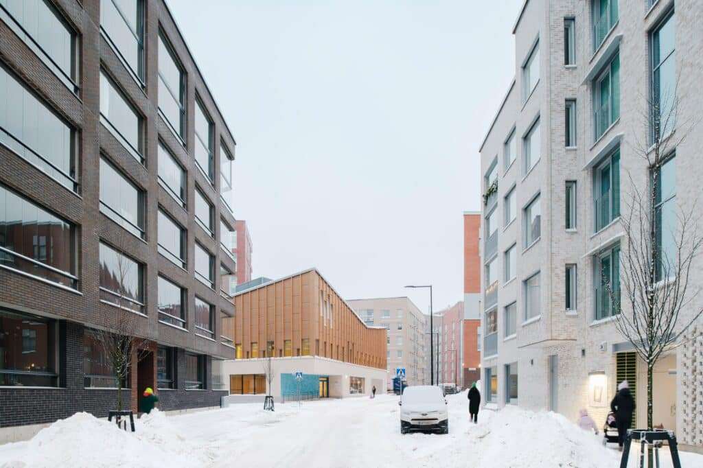 Street view of Postiljooni Daycare Centre in winter, emphasizing its horizontal stone base and the surrounding white-rendered buildings.