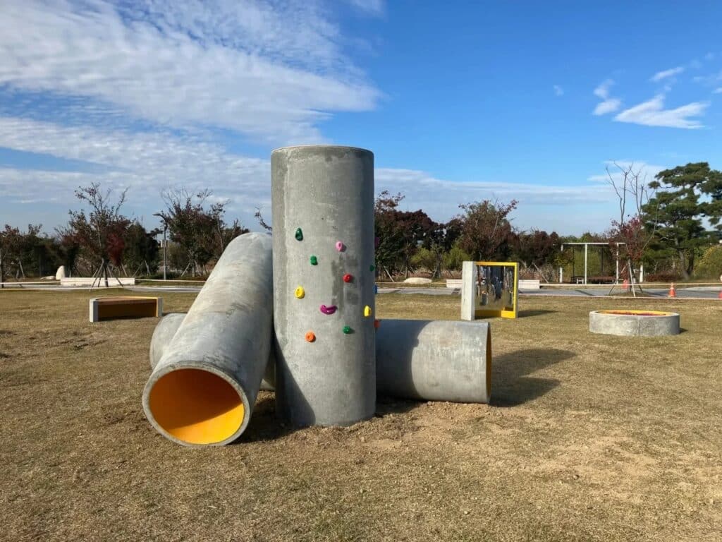 A tall vertical concrete pipe with colorful climbing holds attached to its surface, surrounded by horizontal pipes.
