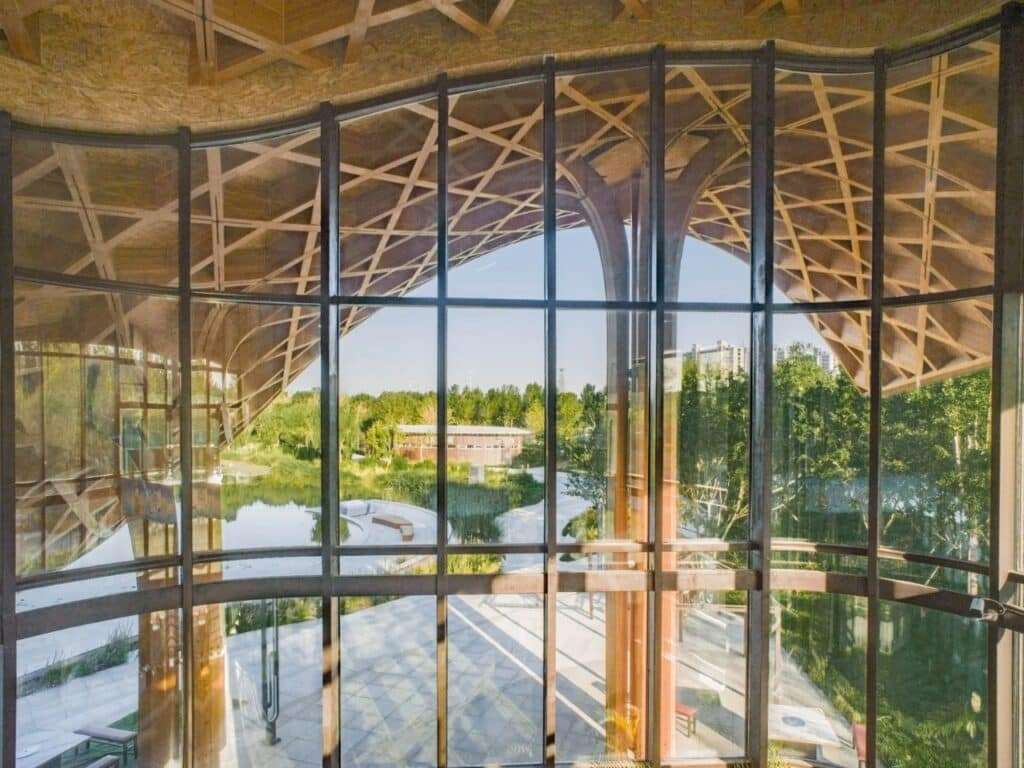 Interior view looking out through a curved glass curtain wall and timber grid shell roof towards the park.