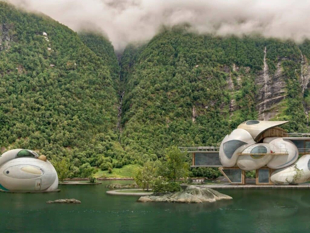 Organic, bubble-like architectural structures floating on a calm lake beneath fog-covered green mountains.