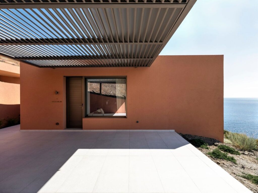 Detail of an earth-toned exterior wall with a minimalist window and a metal slat pergola casting shadows on the terrace.