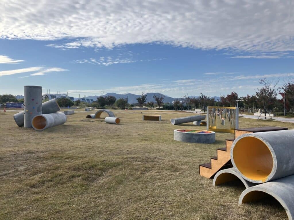 Landscape view of the Concrete Utopia project featuring pipes, wooden stairs, and a curved mirror element in a park setting.
