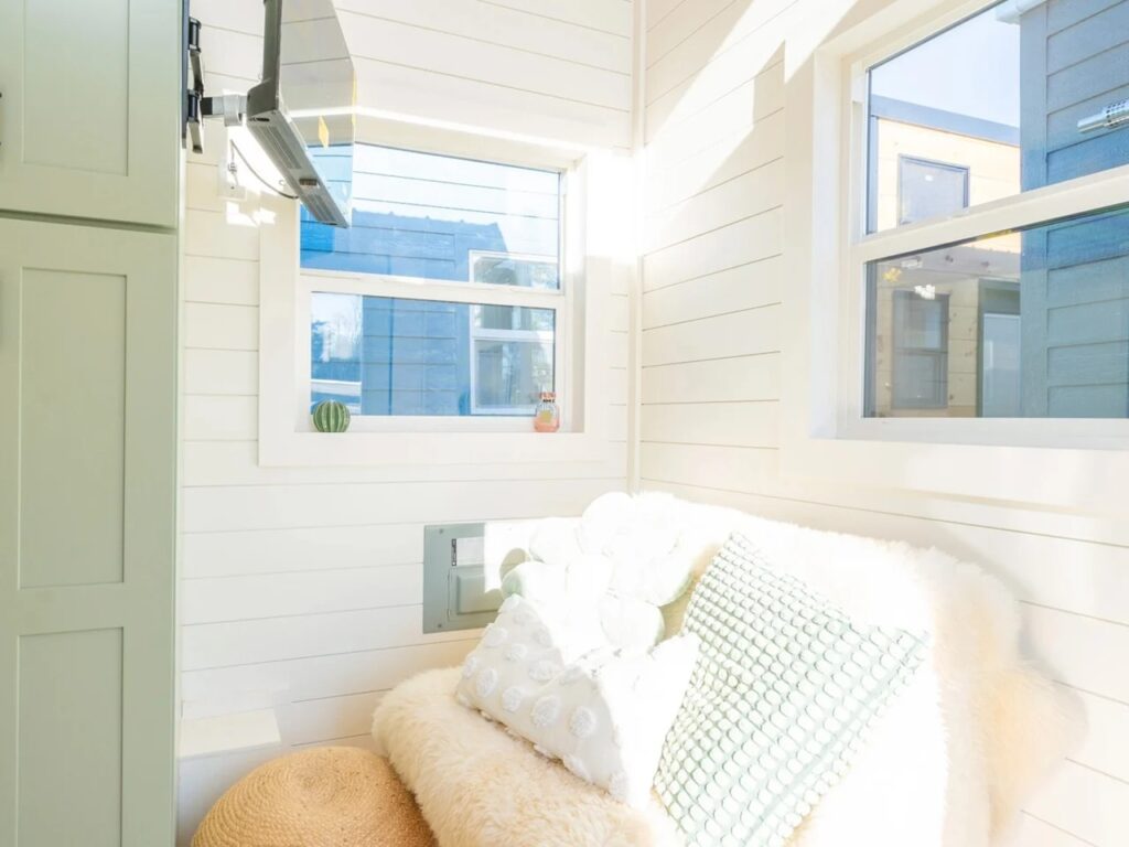 Interior of a tiny house living area with a white fur sofa, green cushions, and a wall-mounted TV near a window.