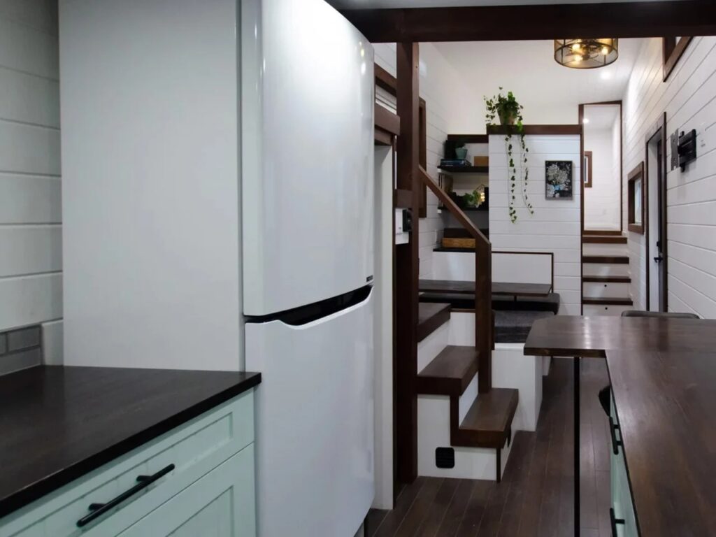 Compact kitchen and hallway in a tiny house featuring a white refrigerator and wooden stairs with storage.