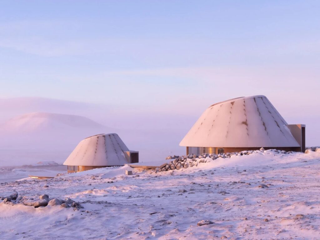Close-up of two architectural cabins with reddish metal panels and aluminum roofs against a hazy volcanic backdrop.