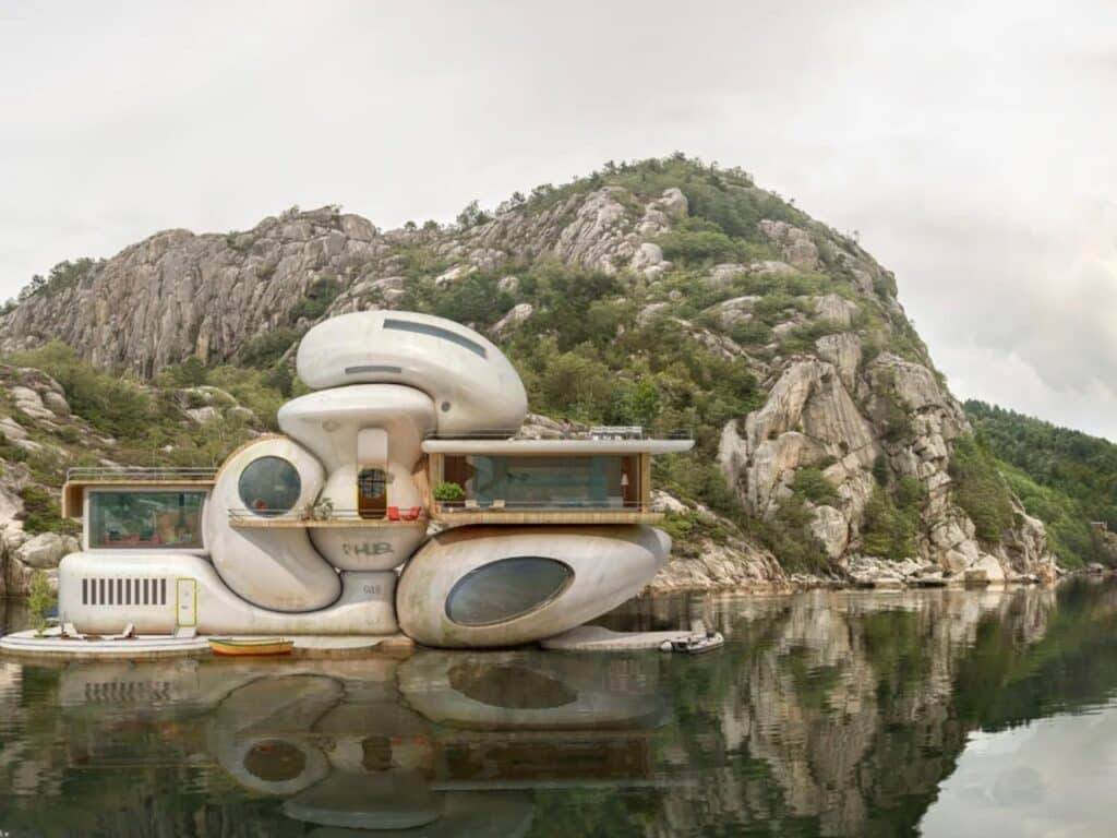 A multi-level futuristic floating home with rounded edges and large porthole windows reflecting in still water.