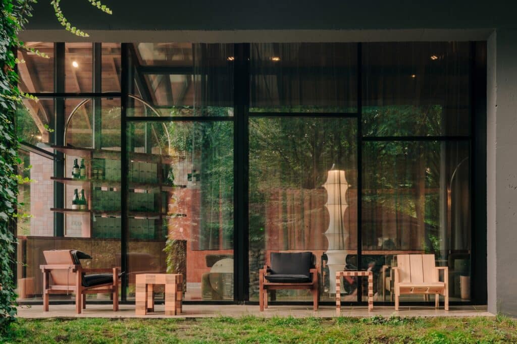 Exterior view of a glass-walled pavilion with wooden lounge chairs and green garden surroundings.