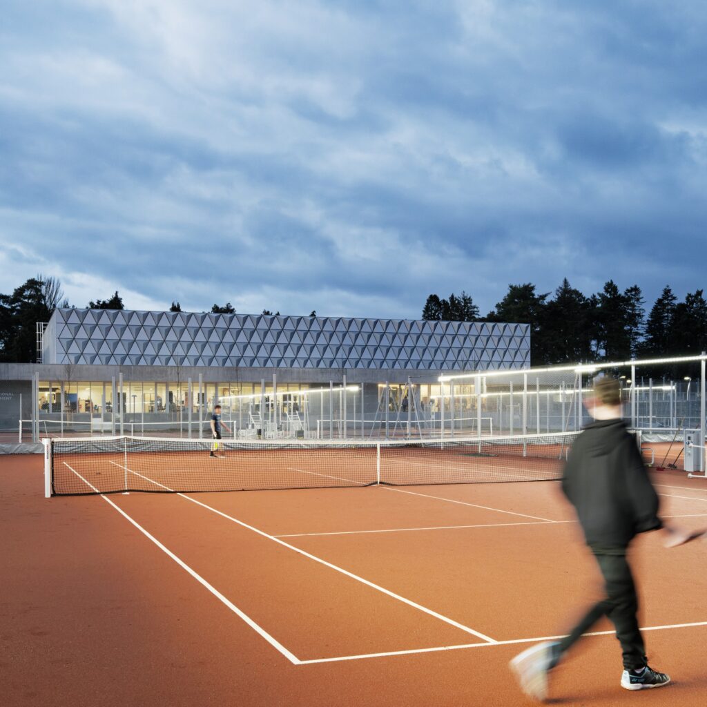 Night view of an outdoor tennis court at Sporting Vichy with the illuminated textured facade of the main hall in the background.