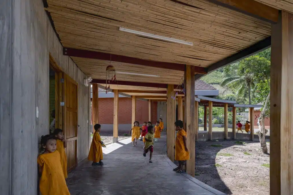 Perspective view of a long wooden corridor with bamboo roofing and children running between classrooms at Sonomoro School.