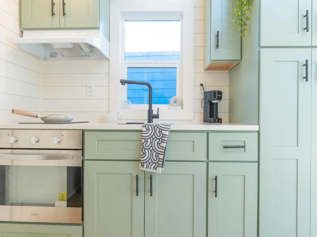 Modern tiny house kitchen with sage green cabinets, white countertops, a black faucet, and an integrated oven.