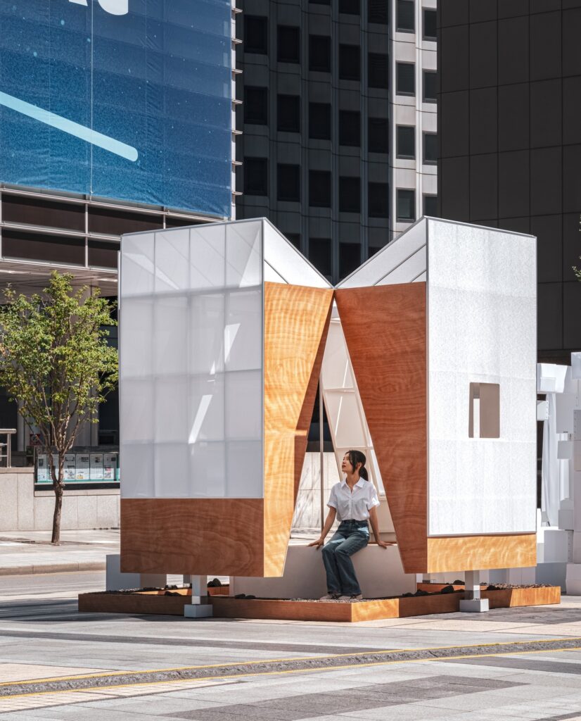 A woman sits on a central bench inside the TEUM pavilion, looking upward at the sunlight filtering through the structure&rsquo;s white and wood walls.