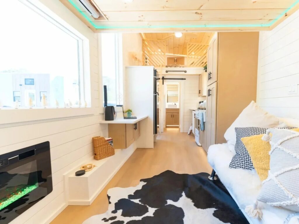 Open living area of a tiny home with a panoramic window, electric fireplace, and floating desk.