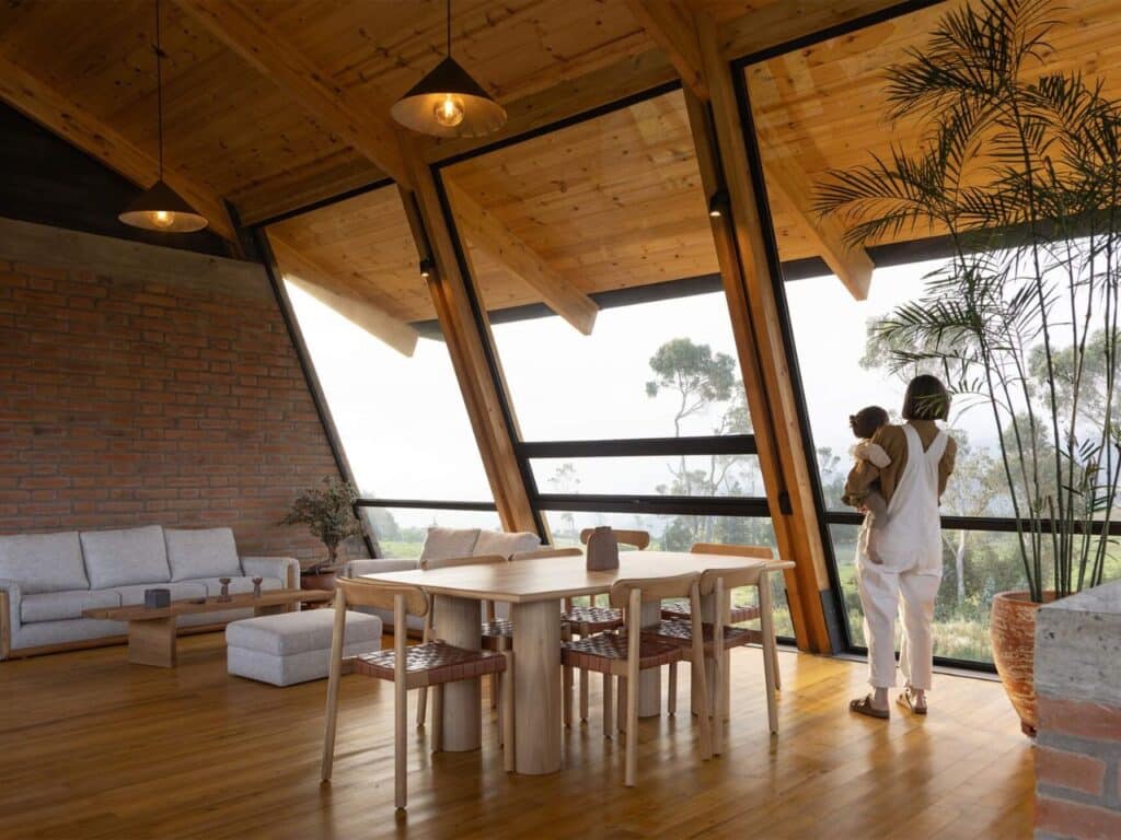 Open plan living room in La Miradora with timber ceilings, large slanted windows, and a person holding a child looking at the view.
