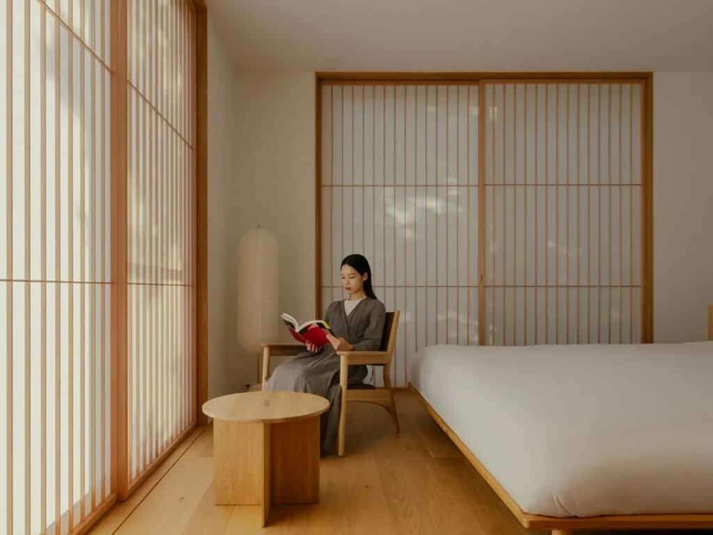 Minimalist bedroom with shoji-style sliding screens and a person reading a book in a wooden chair.