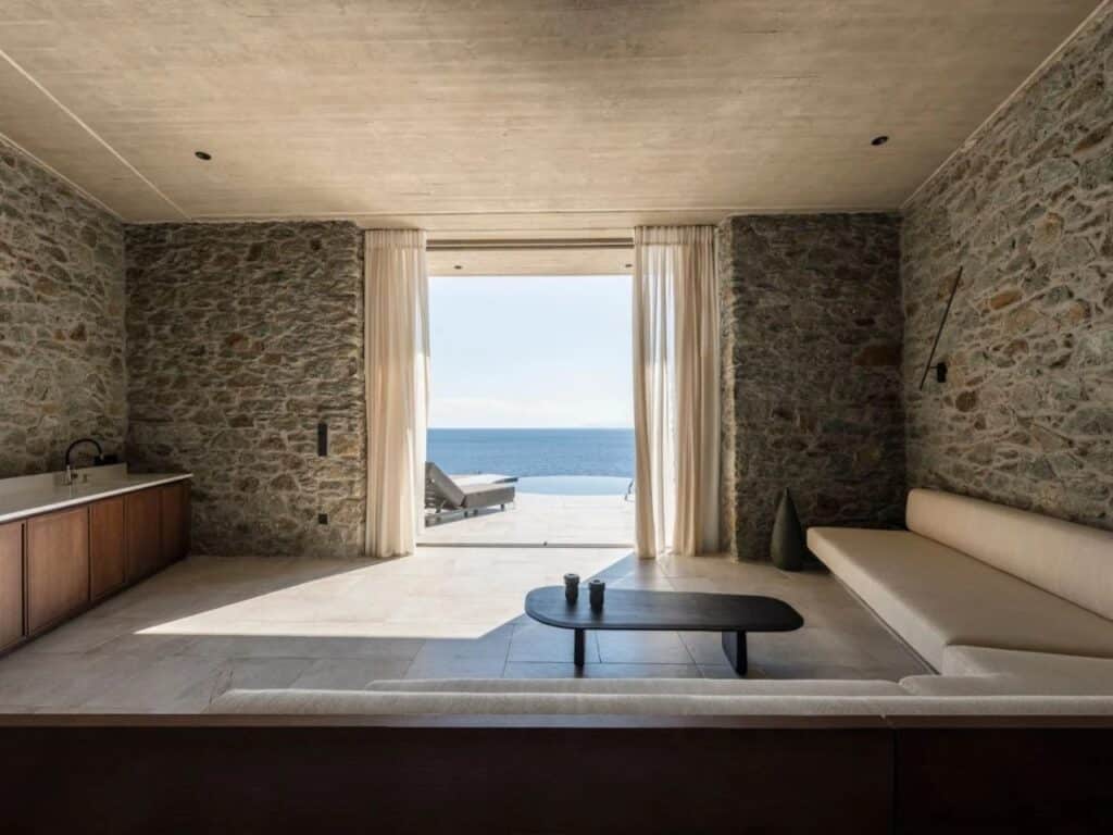 Spacious living room with exposed stone walls, minimalist furniture, and a direct view of the sea through a large opening.