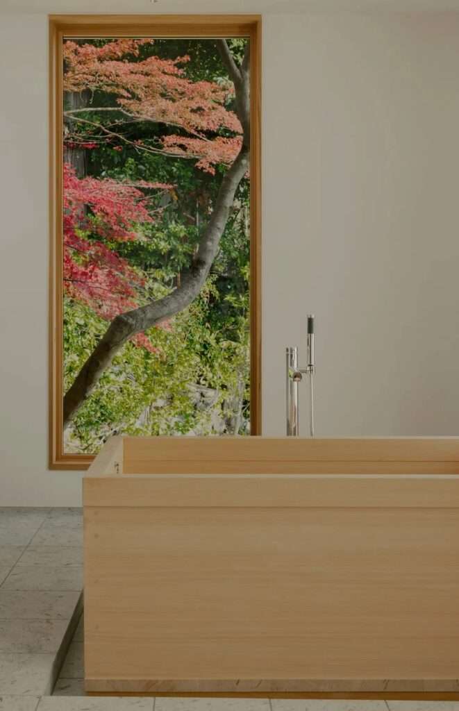 A wooden Hinoki bathtub in a modern bathroom with a large window framing vibrant autumn leaves.