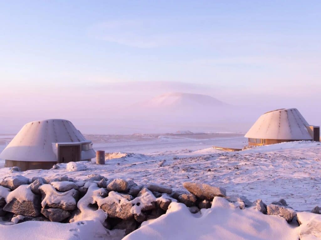 Architectural cabins framed by a traditional dry stone wall in a snow-covered volcanic field.