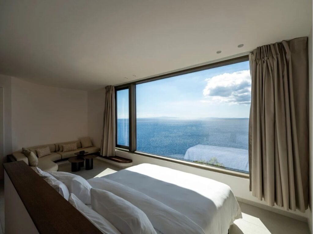 Minimalist bedroom interior at Oline resort with a large panoramic window framing the Aegean Sea and a light-colored lounge area.