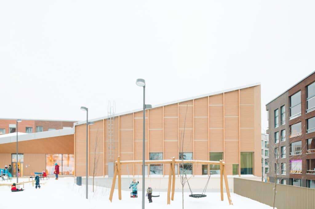 Outdoor playground of Postiljooni Daycare with children playing in the snow next to the sculptural wooden canopy.