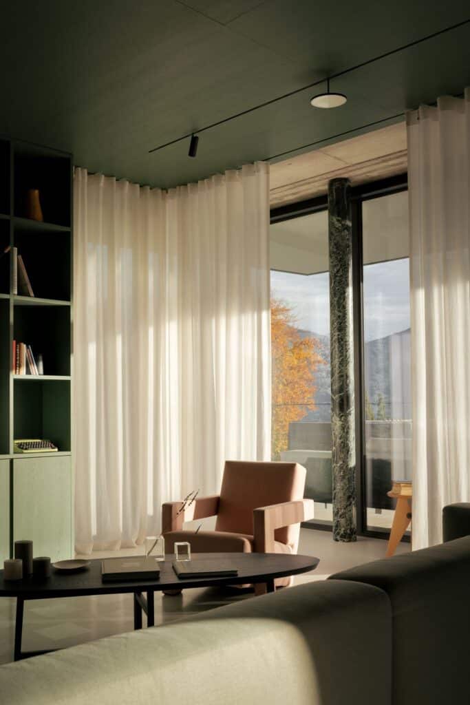 Interior lounge area with sheer wool curtains, a green marble pillar, and a view of autumn trees at Lake Como.