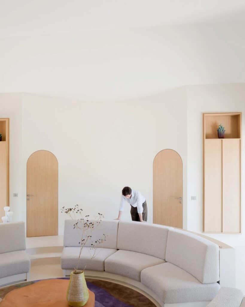 A man leaning on the curved sofa in the minimalist white interior of Seaside House with twin arched doors.