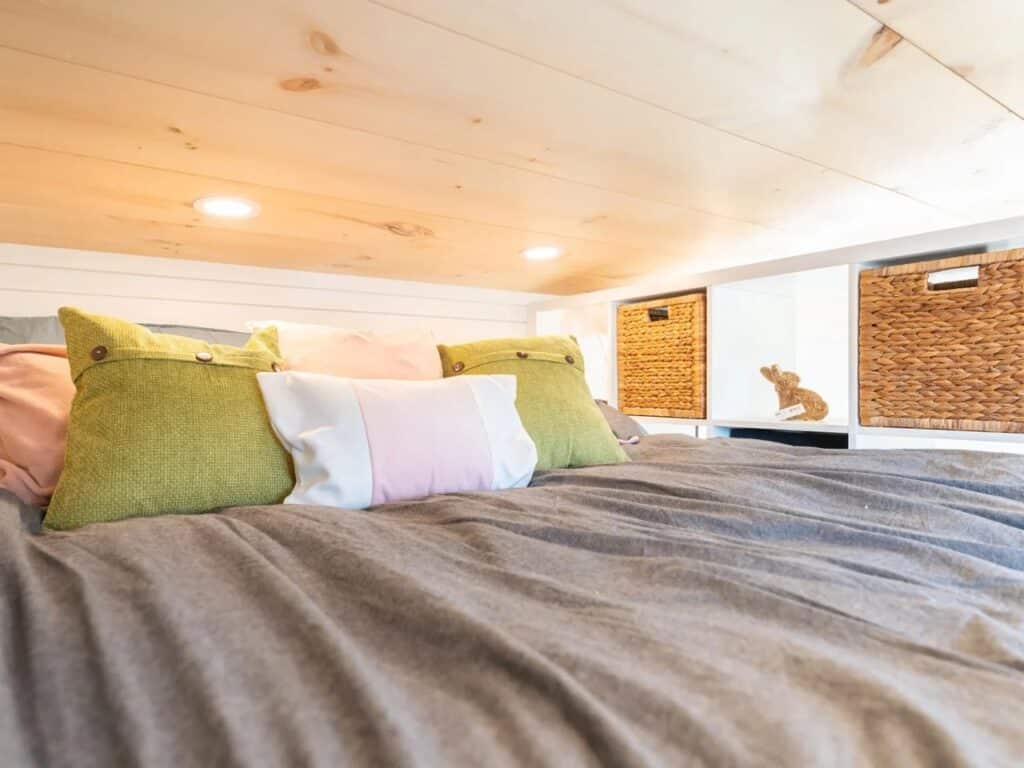 Cozy sleeping loft in a tiny house with a low ceiling, wooden panels, and wicker storage baskets.
