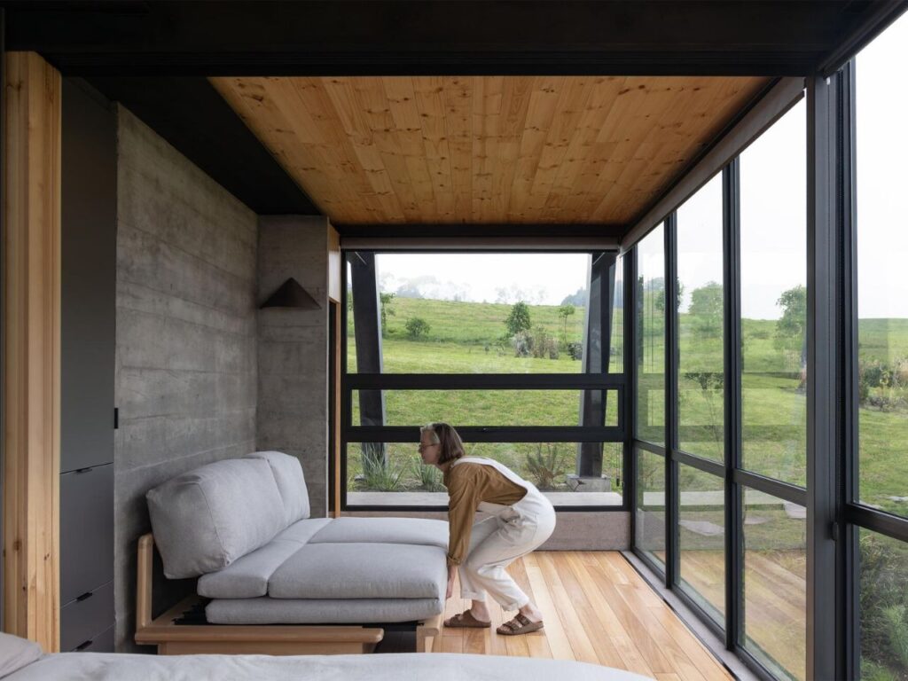 A person adjusting a sofa bed in a compact room with large windows and a timber ceiling in La Miradora.