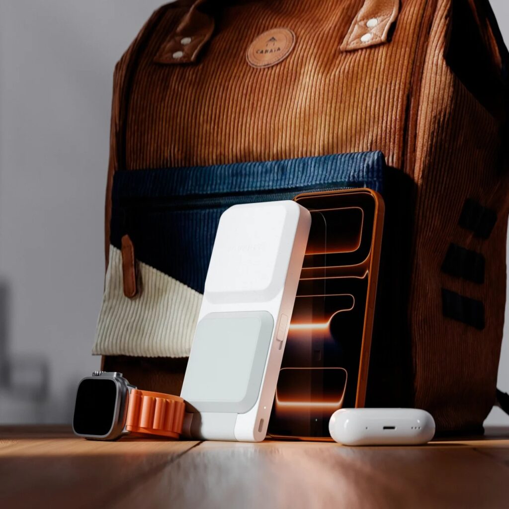 ARIA charger, iPhone, Apple Watch, and AirPods arranged next to a corduroy backpack on a wooden floor in warm sunlight.