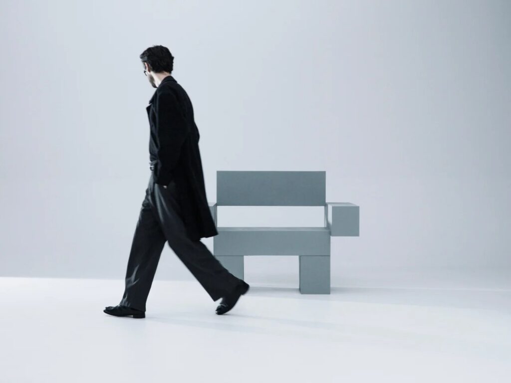 Motion blur of a person walking past the front of THE OBJECT 01 chair in a minimalist white studio.