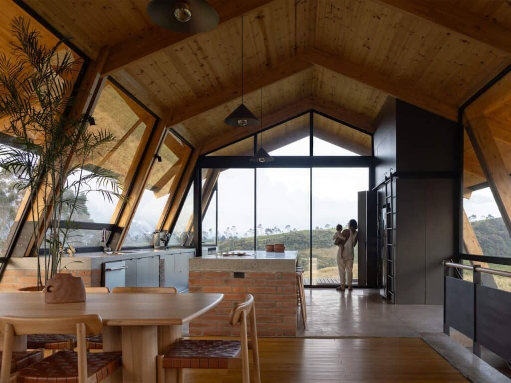 Interior of La Miradora house featuring high gabled timber ceilings, an open-plan kitchen with a brick island, and large glass walls overlooking the Ecuadorian landscape.