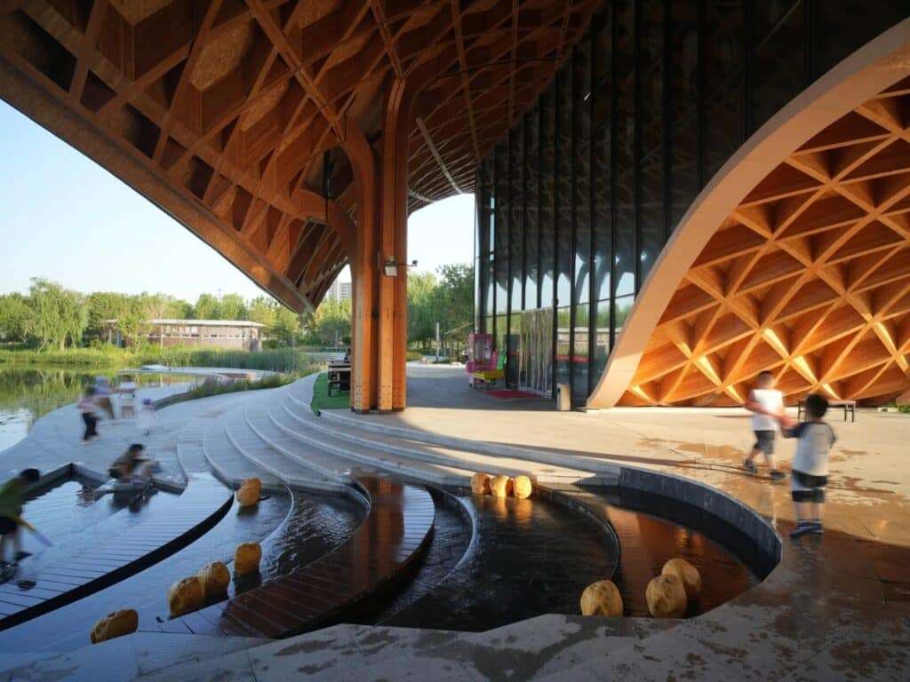 Wide shot of the restaurant's outdoor plaza with children playing near water features under the timber canopy.