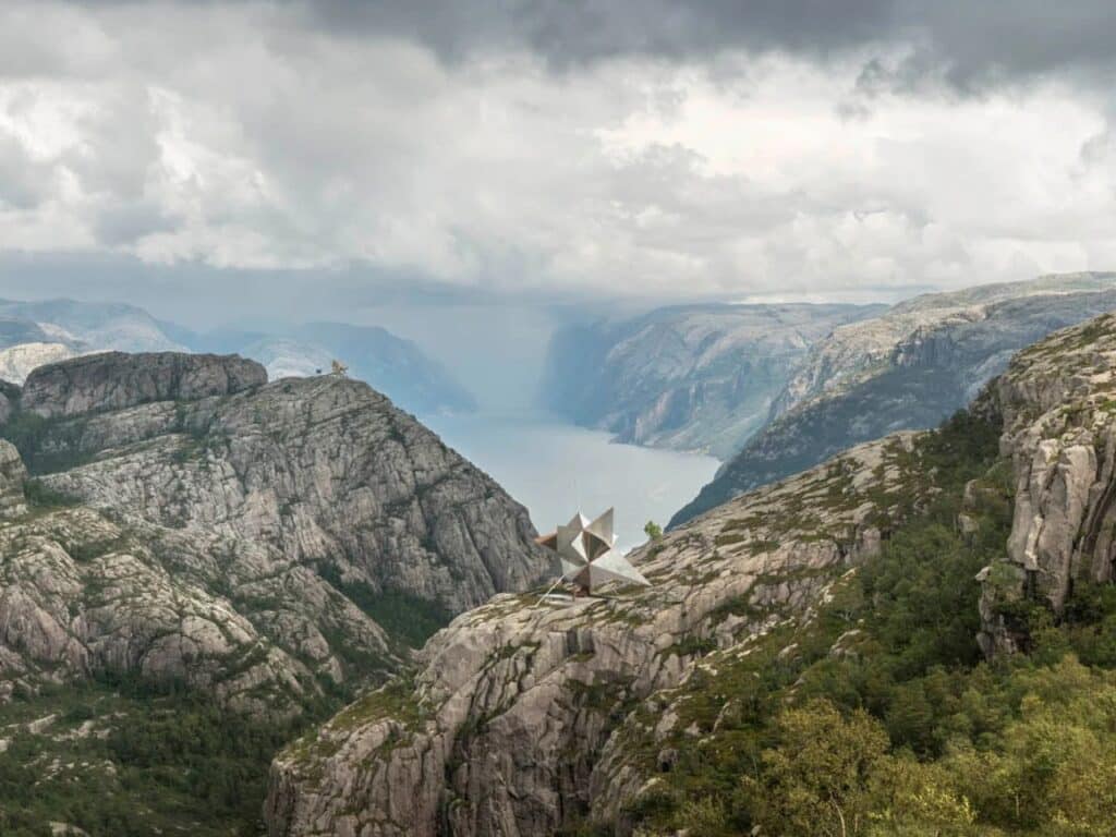 A sharp, star-shaped geometric sculpture perched on a high rocky cliff overlooking a Norwegian fjord under a dramatic cloudy sky.