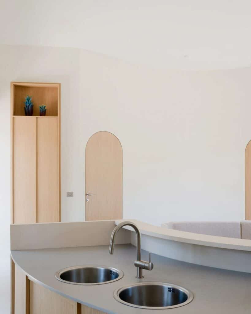 Close-up of a modern minimalist kitchen island with a stainless steel double sink and an arched wooden door in the background.