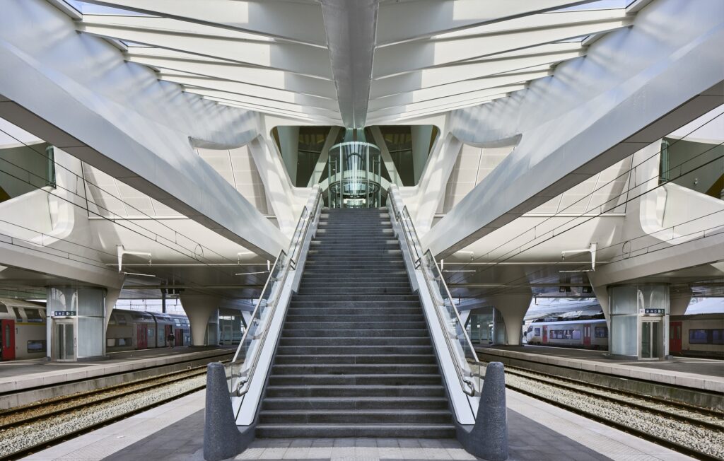 Symmetrical staircase descending to the railway platforms at Mons Station, framed by white steel structural elements.