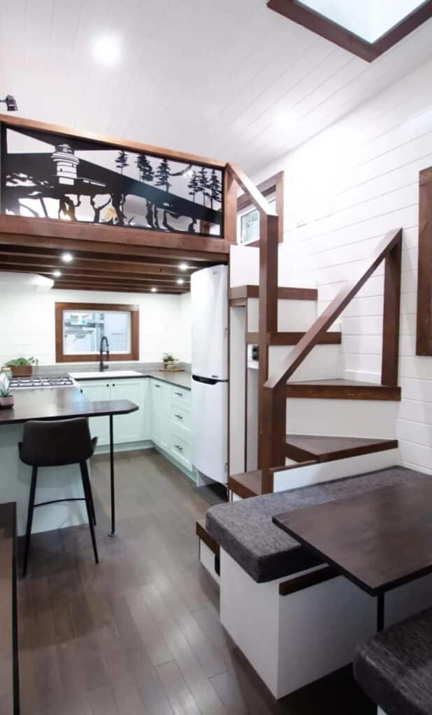 A vertical interior wide-angle shot of the Starling tiny home, featuring a convertible dining nook, a mint-colored U-shaped kitchen, and a primary loft bedroom with a decorative metal railing.