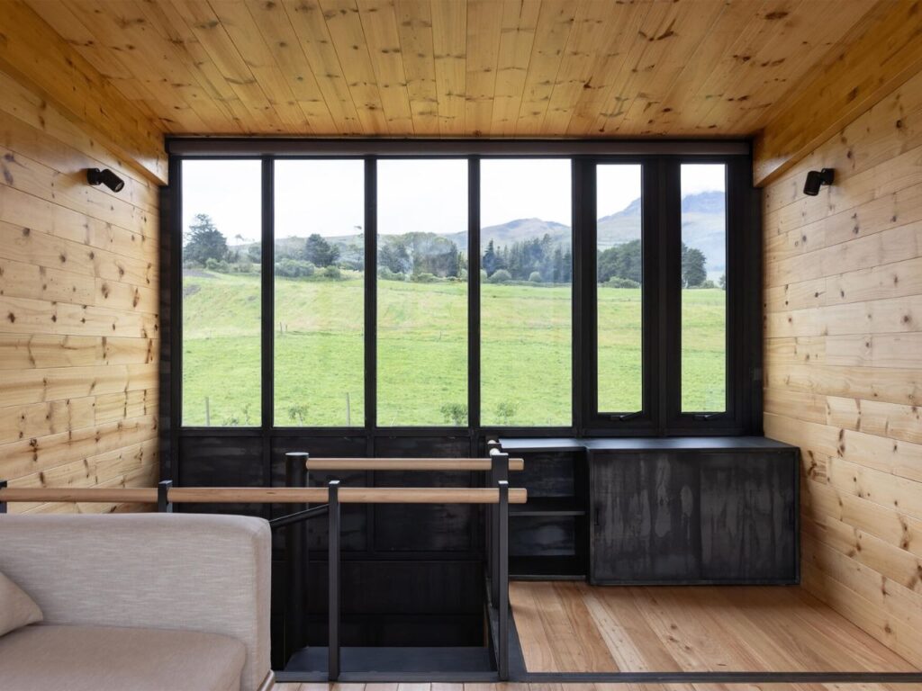 Interior mezzanine or loft space in La Miradora with timber-clad walls, black metal railings, and a wide window facing green hills.
