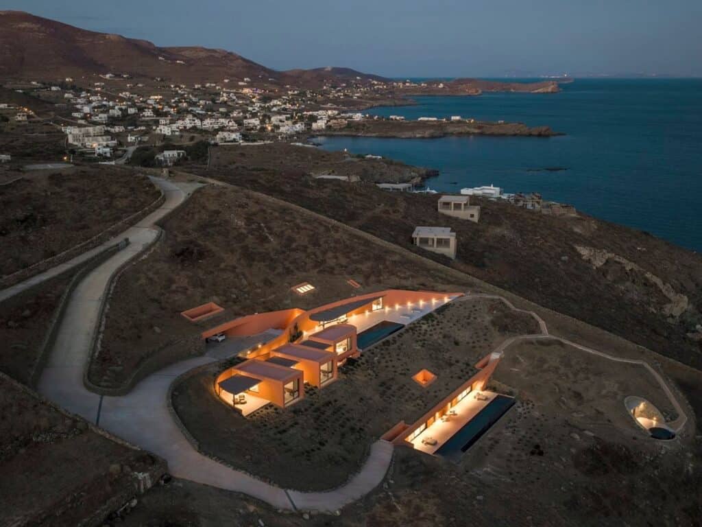 Wide aerial dusk view of the Oline resort project on a Syros hillside, showing the glowing units and the coastline in the background.