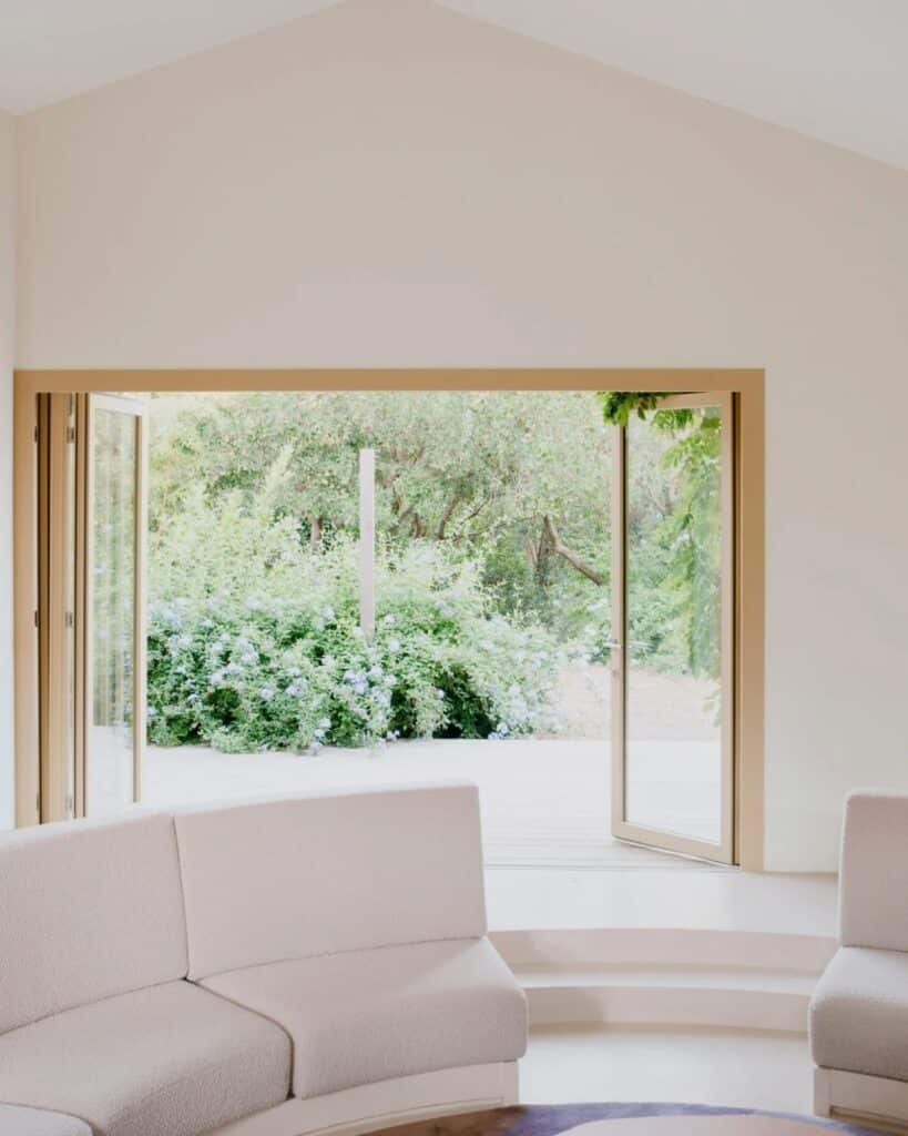 Wide open glass doors in the Seaside House living room looking out onto a sunlit garden and lush shrubs.