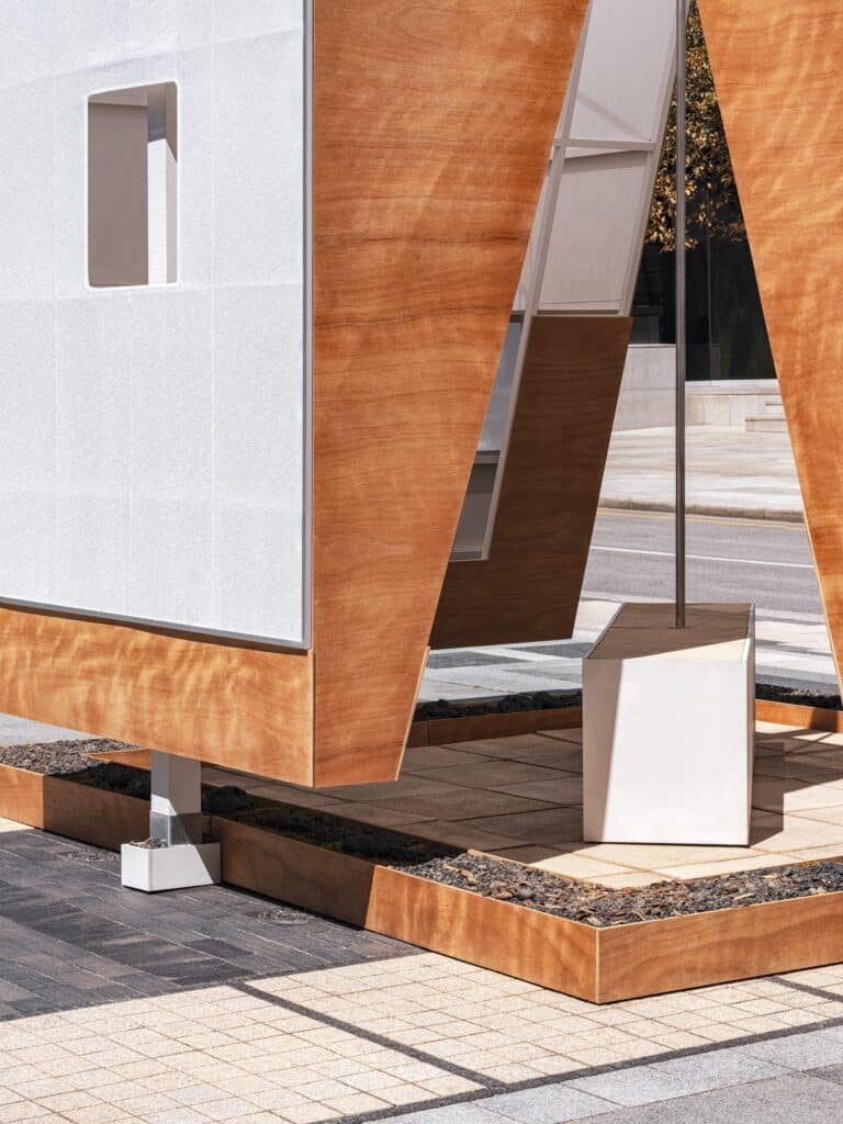 A close-up detail of the base of the TEUM pavilion showing wooden panels resting on a gravel-filled foundation next to a tiled sidewalk.