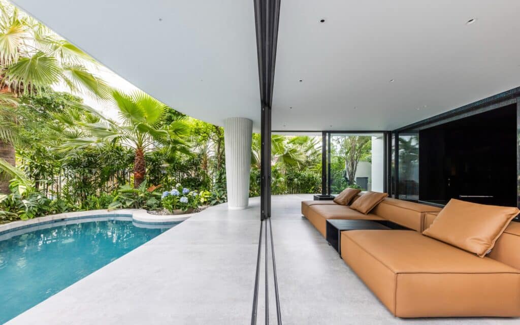 Open-plan living room with a tan leather sofa next to an outdoor swimming pool and tropical plants.