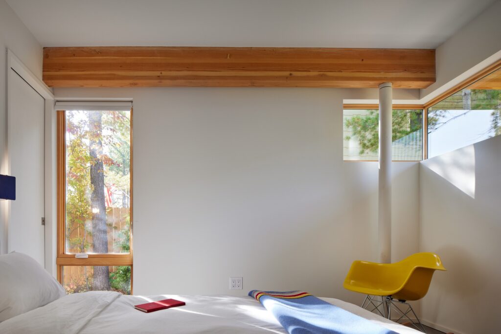 Bright bedroom interior with a large wooden beam, white walls, and a yellow Eames-style chair near a window.