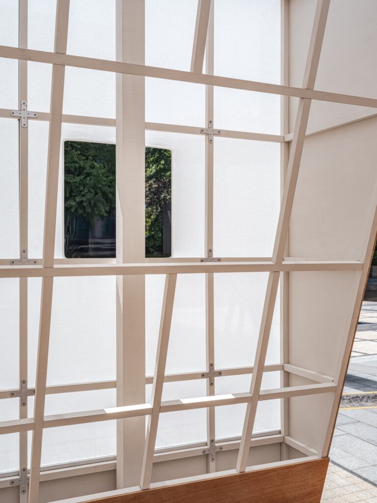 Interior detail of a white translucent gridded wall with a small rectangular opening framing green tree leaves outside.