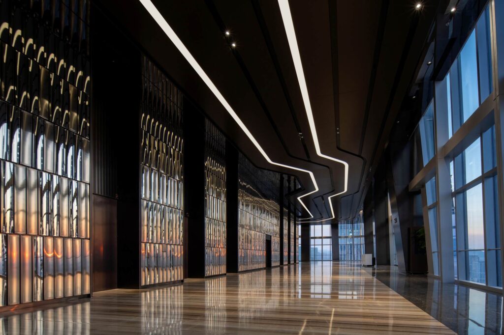 A spacious interior lobby with a reflective floor, textured mirrored walls, and a zigzag ceiling light leading toward a panoramic window view at sunset.