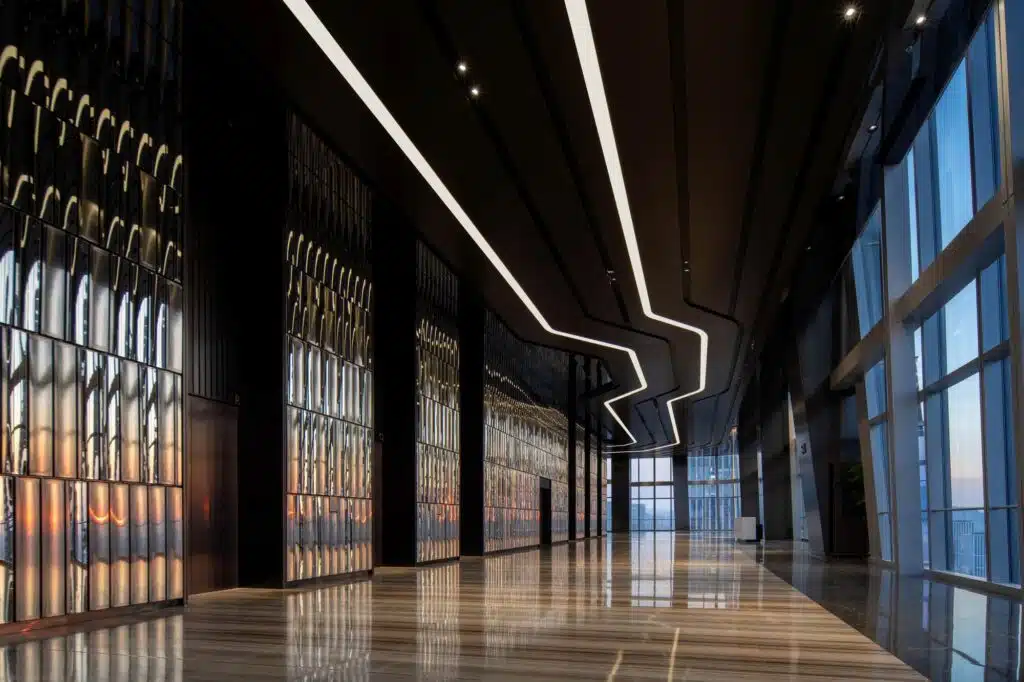 A spacious interior lobby with a reflective floor, textured mirrored walls, and a zigzag ceiling light leading toward a panoramic window view at sunset.