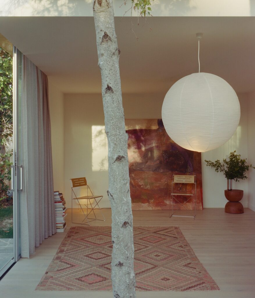 Interior view through a birch tree trunk showing a minimalist room with a large spherical lamp and an abstract painting.