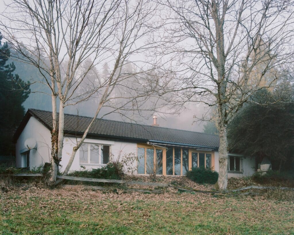 Exterior view of the 1983 building showing its low profile, tiled roof, and the new large wooden windows integrated into the white facade amidst a forest setting.