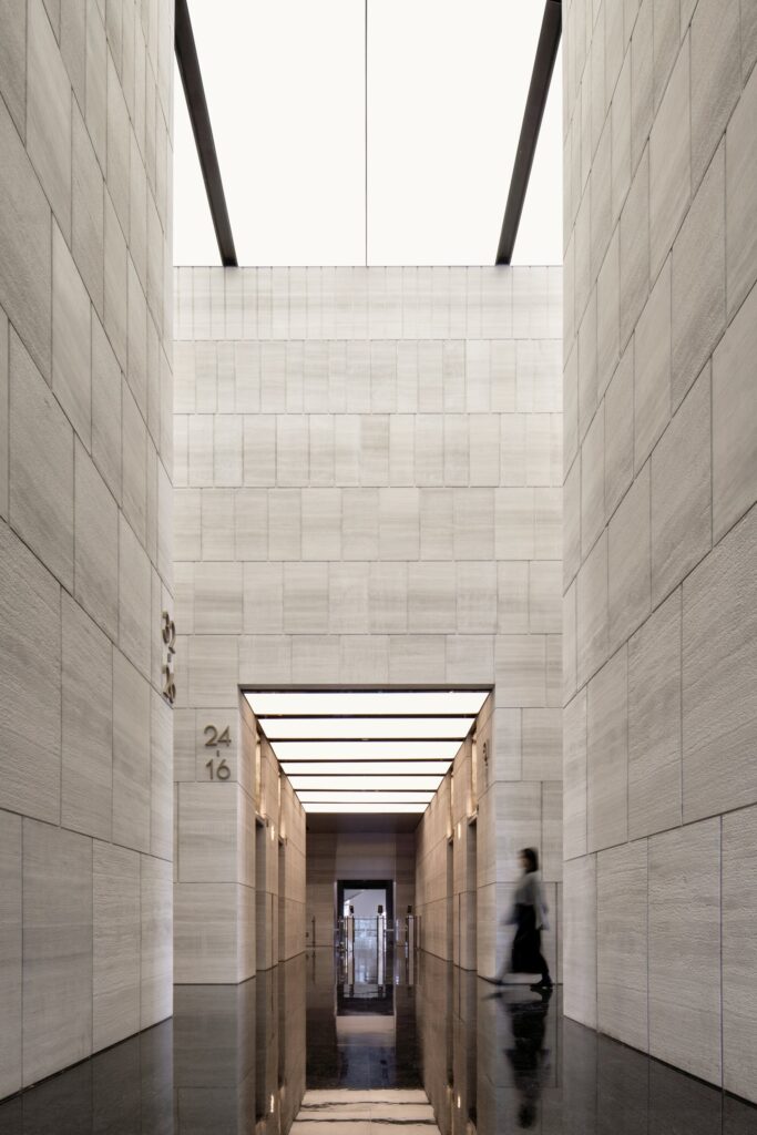 A low-angle perspective of a narrow stone-clad atrium in Jinan Tower, leading towards a bright, luminous ceiling with a person walking in the background.