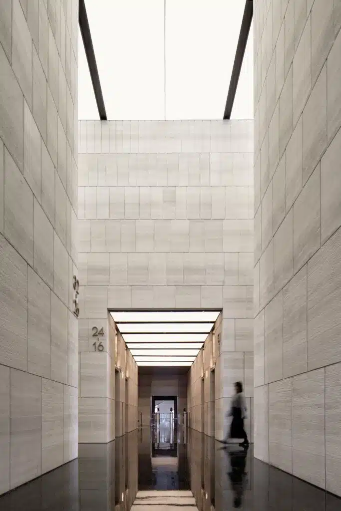A low-angle perspective of a narrow stone-clad atrium in Jinan Tower, leading towards a bright, luminous ceiling with a person walking in the background.